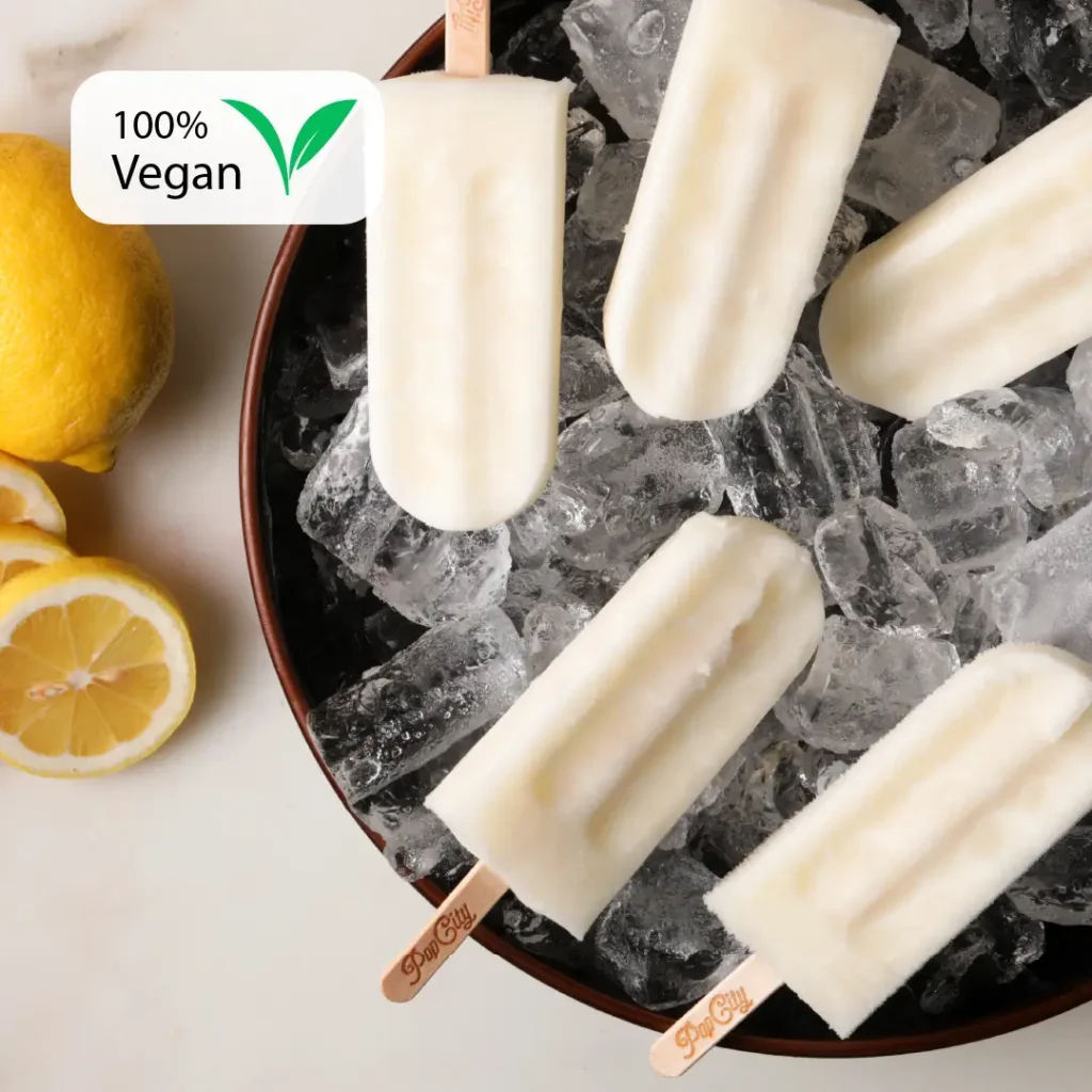 White lemon-flavored popsicles on a bed of ice in a bowl, surrounded by fresh lemons and lemon slices, with a "100% Vegan" label in the top left corner and "Pop City" engraved on the sticks.