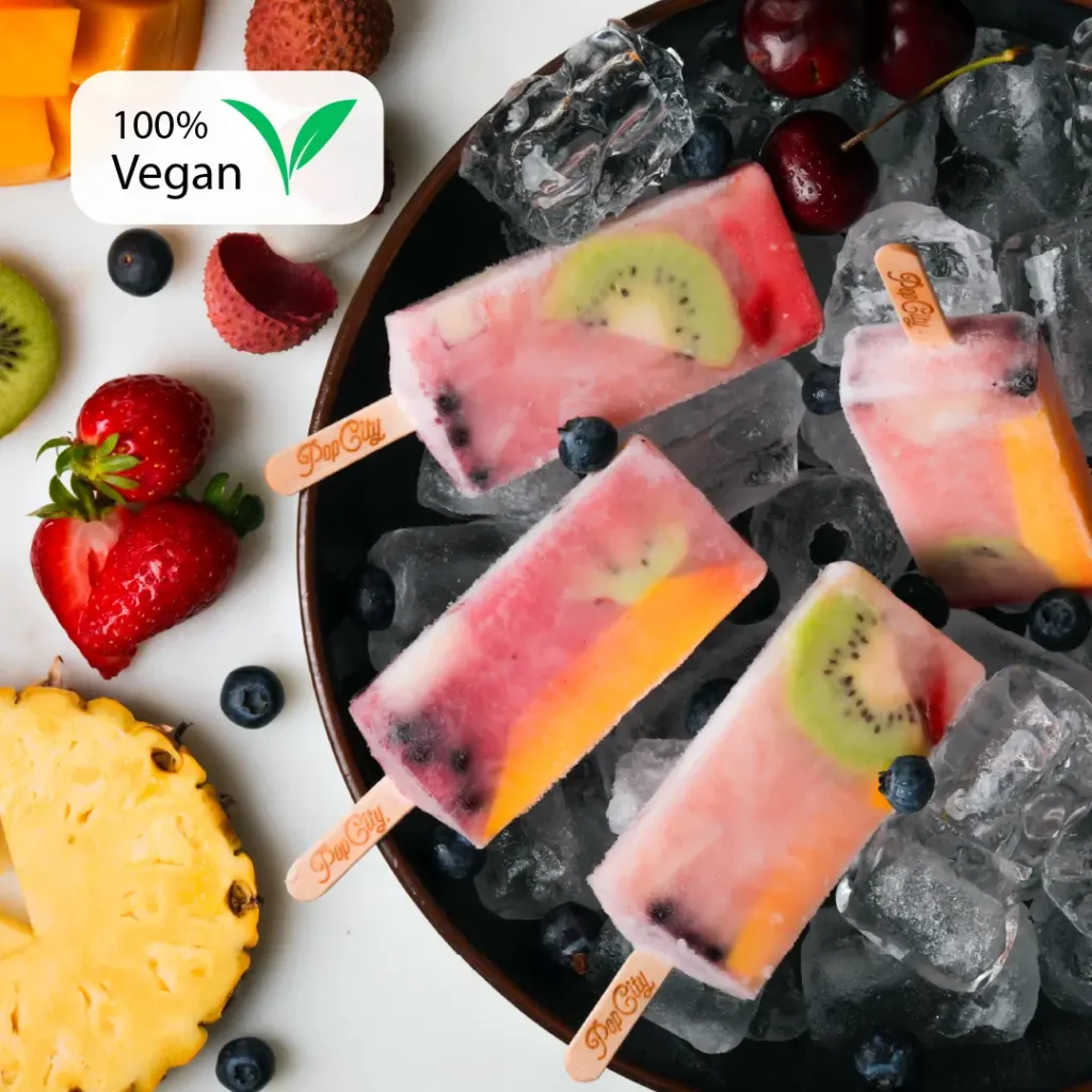 White lychee-flavored popsicles on a bed of ice cubes, surrounded by fresh lychees, strawberries, and blueberries, with a "100% Vegan" label in the top left corner.