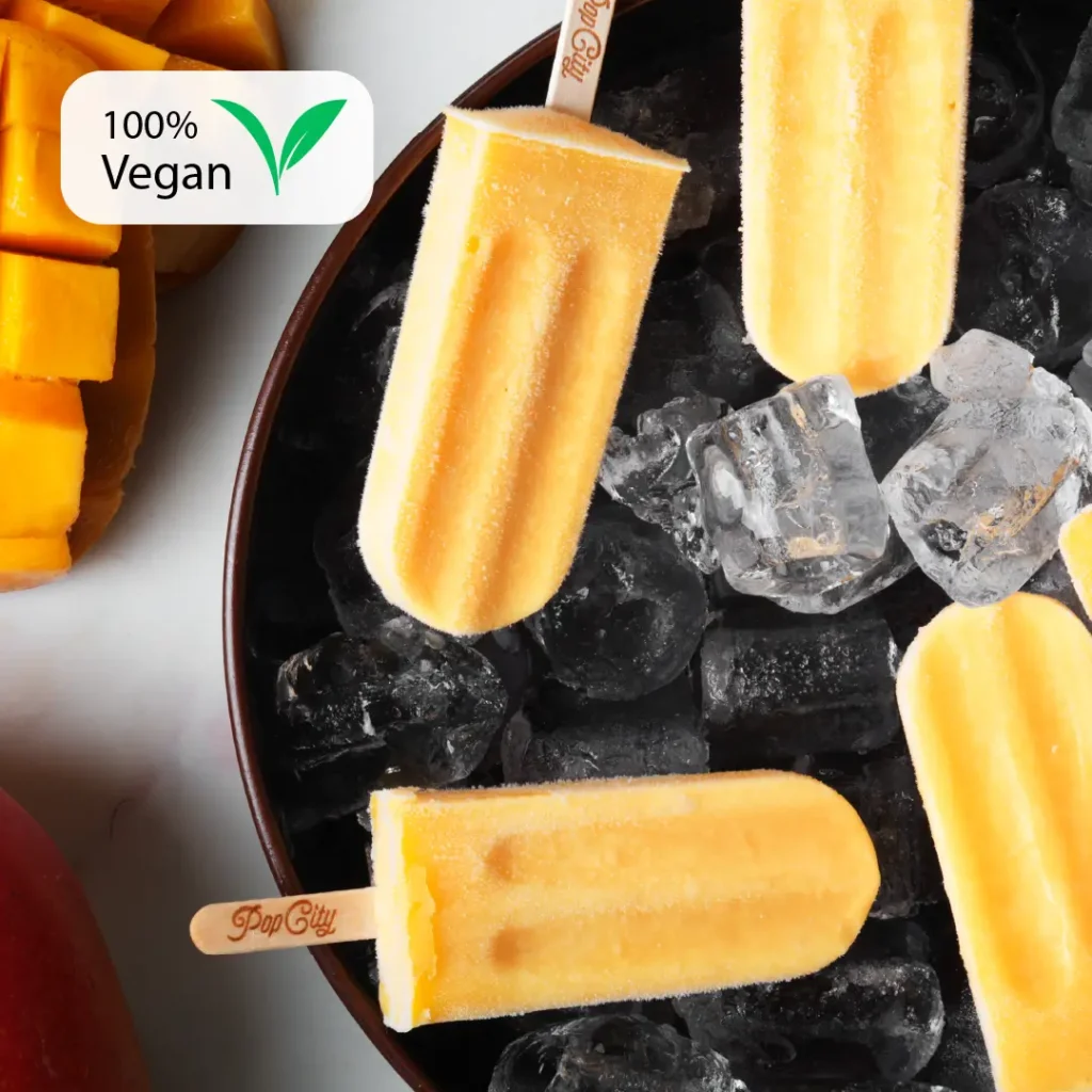 Bright yellow-orange mango-flavored popsicles on a bed of ice, with "Pop City" engraved on the sticks, and a "100% Vegan" label in the top left corner, surrounded by cut mango pieces.