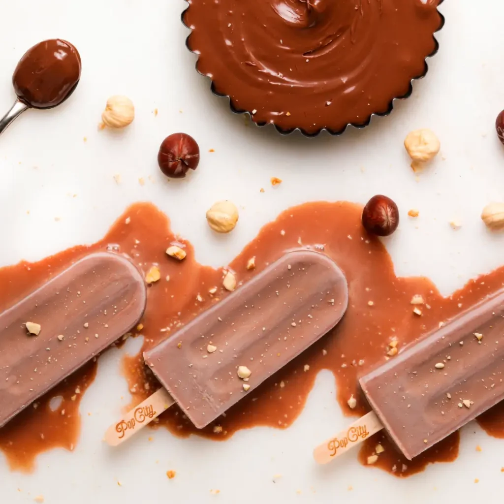 Three chocolate popsicles on a white surface, partially melted with chocolate sauce spread around them, with scattered hazelnuts and a spoon with chocolate sauce nearby.