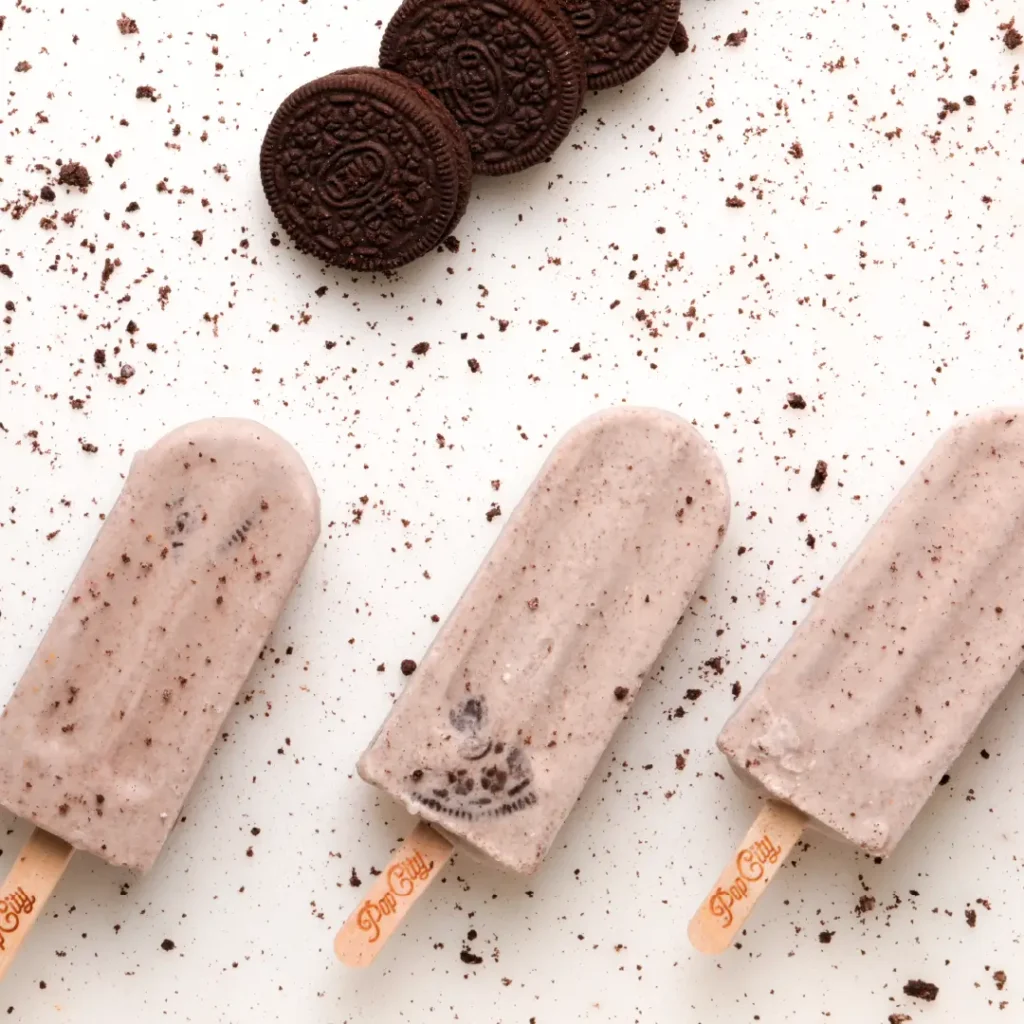 Cookies and cream flavored popsicles on wooden sticks engraved with "Pop City," surrounded by chocolate sandwich cookies and cookie crumbs on a white surface.