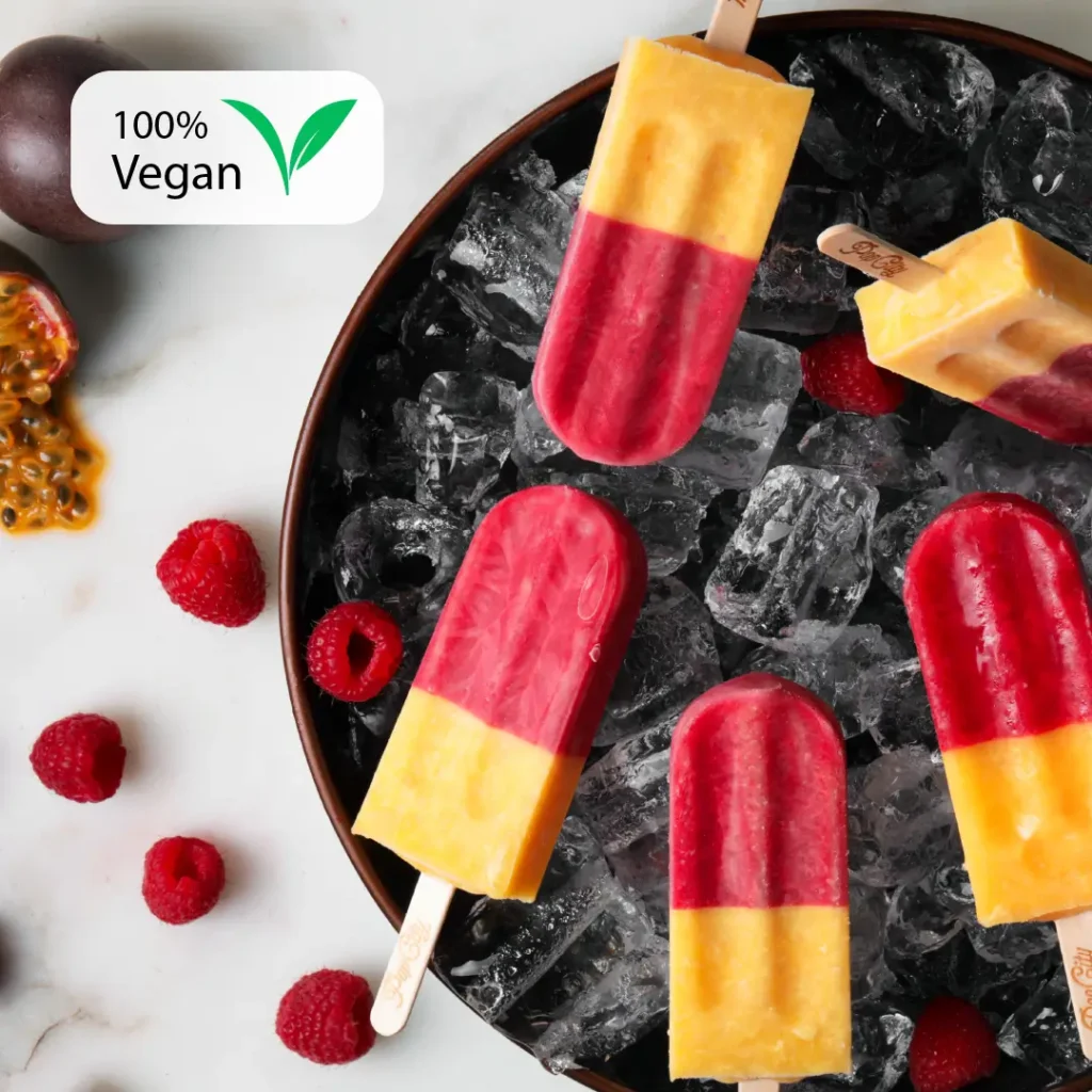 Raspberry and passion fruit flavored popsicles, two-toned with a reddish top and a yellow bottom, on a bed of ice, surrounded by fresh raspberries and a halved passion fruit, with a "100% Vegan" label in the top left corner.