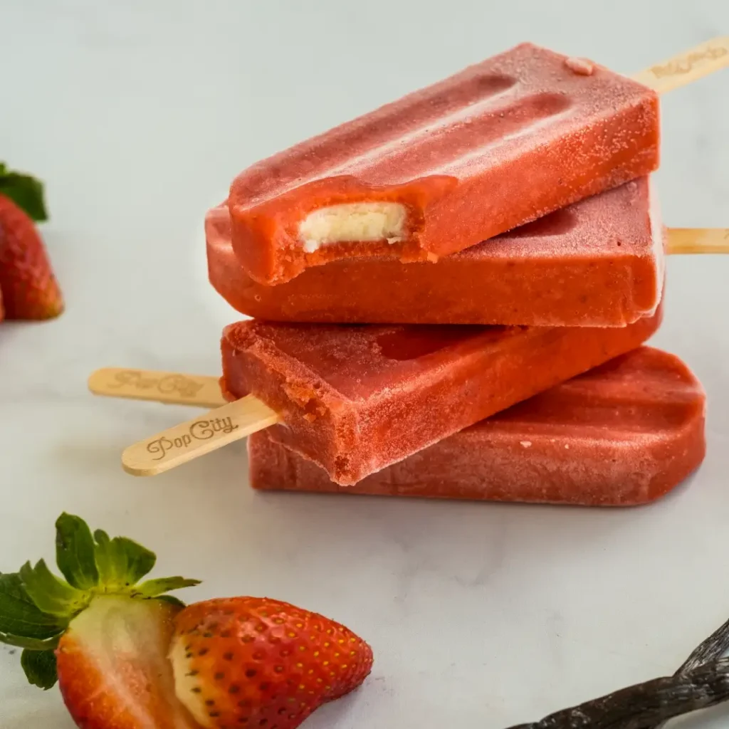 Three strawberry-flavored popsicles with creamy filling, stacked on each other, with fresh strawberries and a vanilla bean in the background.