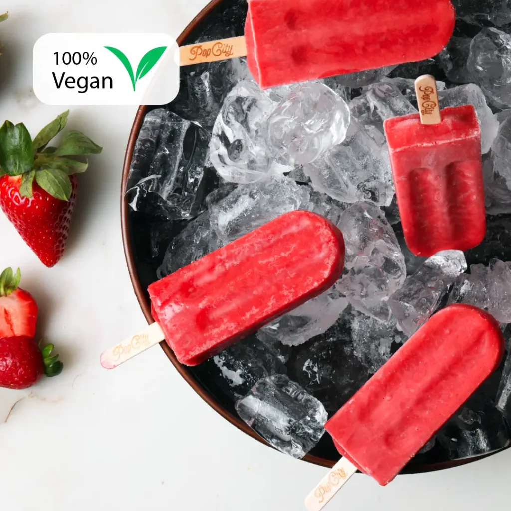 A bowl filled with ice and several red strawberry-flavored popsicles with wooden sticks that have "Pop City" written on them, and fresh strawberries placed next to the bowl.