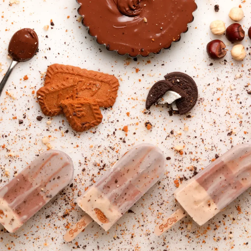 An assortment of dessert items, including layered popsicles, chocolate-filled tarts, cookies, hazelnuts, and chocolate spread, arranged on a white surface.