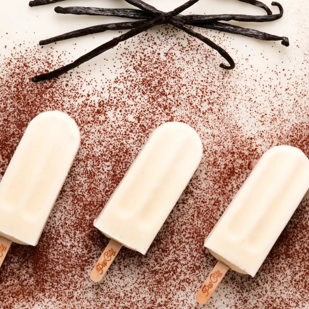 Three vanilla popsicles dusted with cocoa powder, arranged alongside vanilla beans, with "Pop City" text on the sticks.