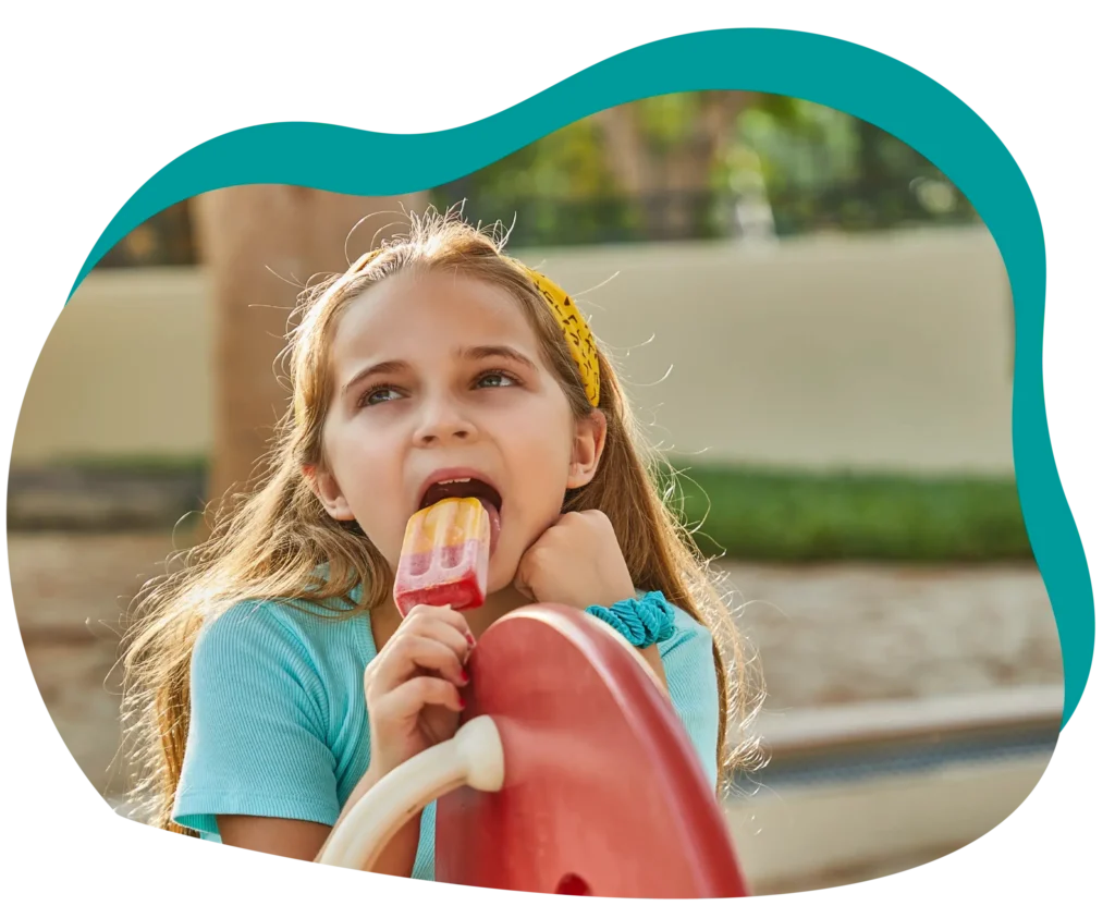 A child with long hair, wearing a blue shirt and a yellow headband, holding a red popsicle from Popcity, with an outdoor playground background.