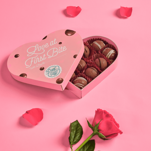 A heart-shaped box of chocolates with the text "Love at First Bite" on the lid, decorated with white lines and partially open, revealing chocolate truffles inside. The background is pink with scattered rose petals and a red rose.