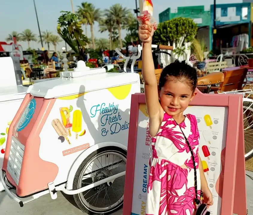 A child holding an ice cream popsicle in front of a Popcity ice cream cart with a sign that reads "Healthy Fresh & Delicious," displayed at an outdoor event with palm trees and seating areas.