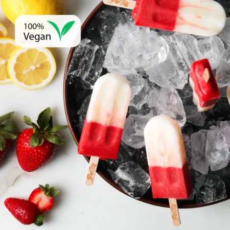 A plate of ice with three strawberry lemon popsicles on sticks, surrounded by fresh strawberries and lemon slices. A label in the top left corner reads "100% Vegan.