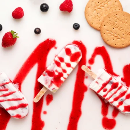 Cheesecake and berry popsicles with a red and white swirl pattern, placed on a white surface with red sauce drizzled around them, surrounded by strawberries, raspberries, blueberries, and two round biscuits.