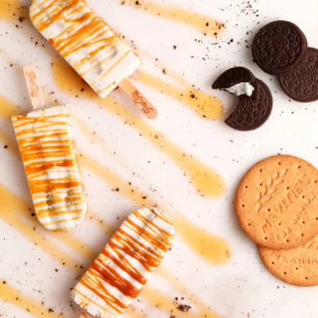 Cheesecake popsicles drizzled with caramel sauce on a white surface, accompanied by Oreo cookies and digestive biscuits with "McVities" embossed on them.