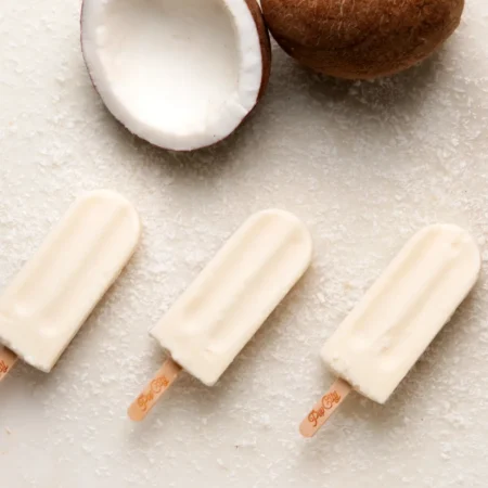 Three creamy coconut popsicles arranged diagonally on a surface sprinkled with shredded coconut, with a halved coconut in the background displaying white coconut flesh and brown outer shell.