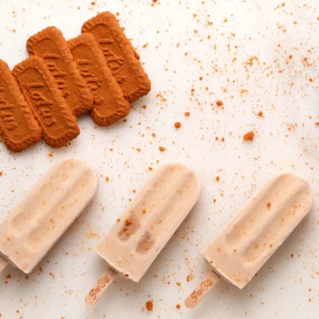 Three creamy popsicles on a white surface with scattered Lotus Biscoff cookies and crumbs, with "Pop City" engraved on the sticks.