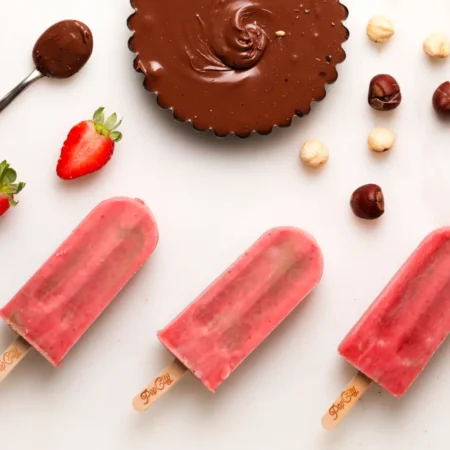 Three strawberry-flavored popsicles arranged horizontally on a white surface, with a tart filled with Nutella, a spoon with Nutella, a halved strawberry, and hazelnuts scattered around.