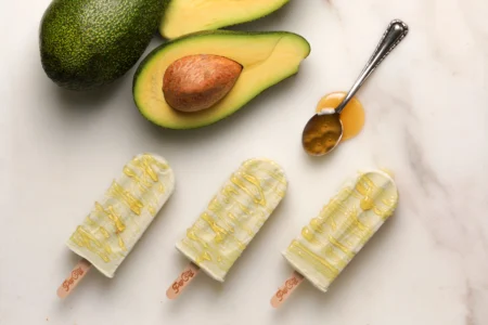 Three avocado popsicles drizzled with honey, surrounded by a whole avocado, a halved avocado with the pit, and a spoon with honey, labeled 'PopCity'