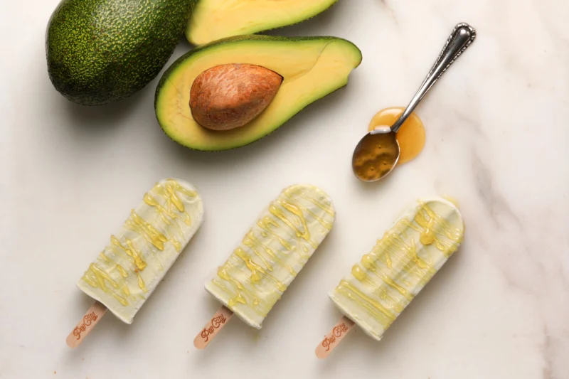 Three avocado popsicles drizzled with honey, surrounded by a whole avocado, a halved avocado with the pit, and a spoon with honey, labeled 'PopCity'