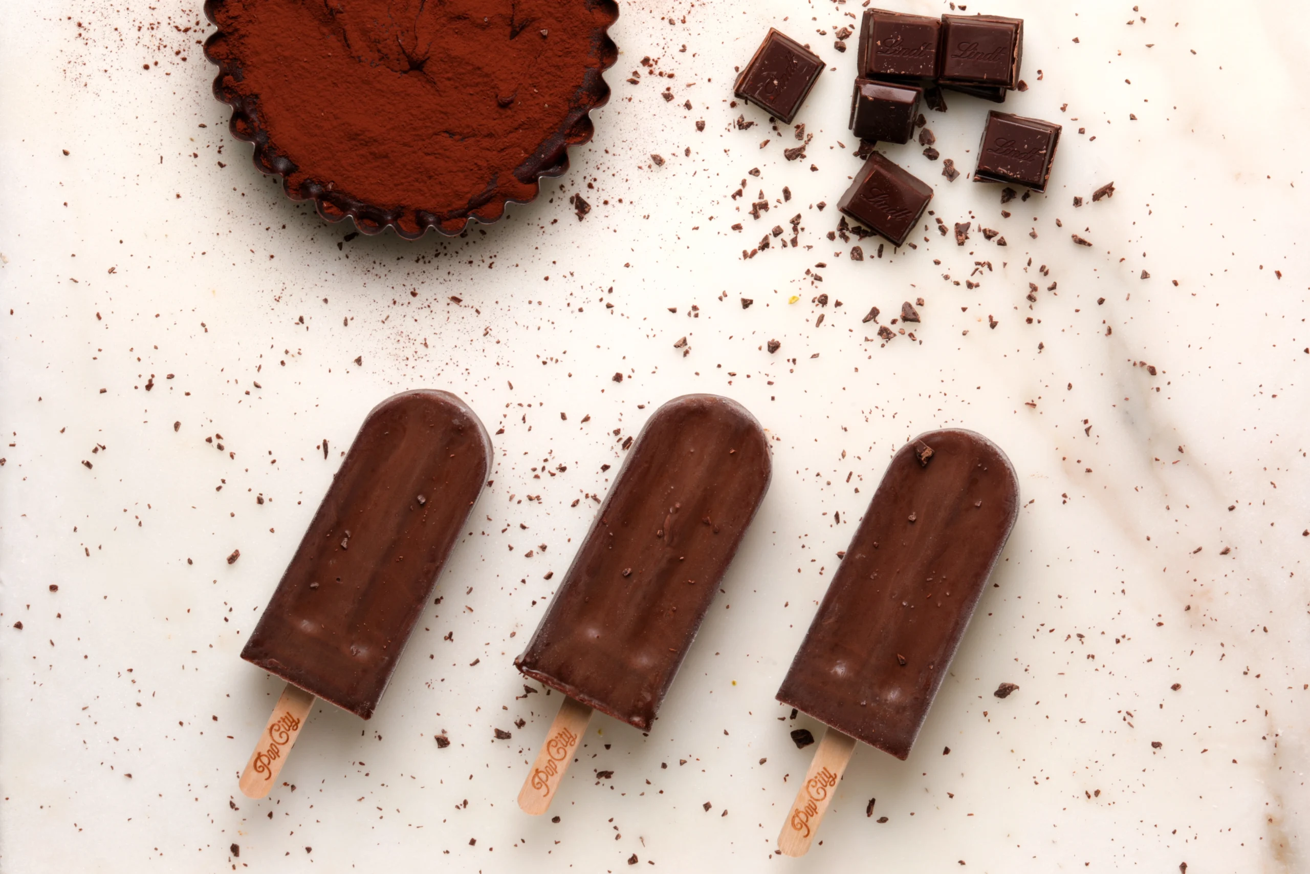 Three decadent dark chocolate popsicles on a white surface, surrounded by chocolate shavings, a cocoa-dusted tart, and dark chocolate squares, labeled 'PopCity'