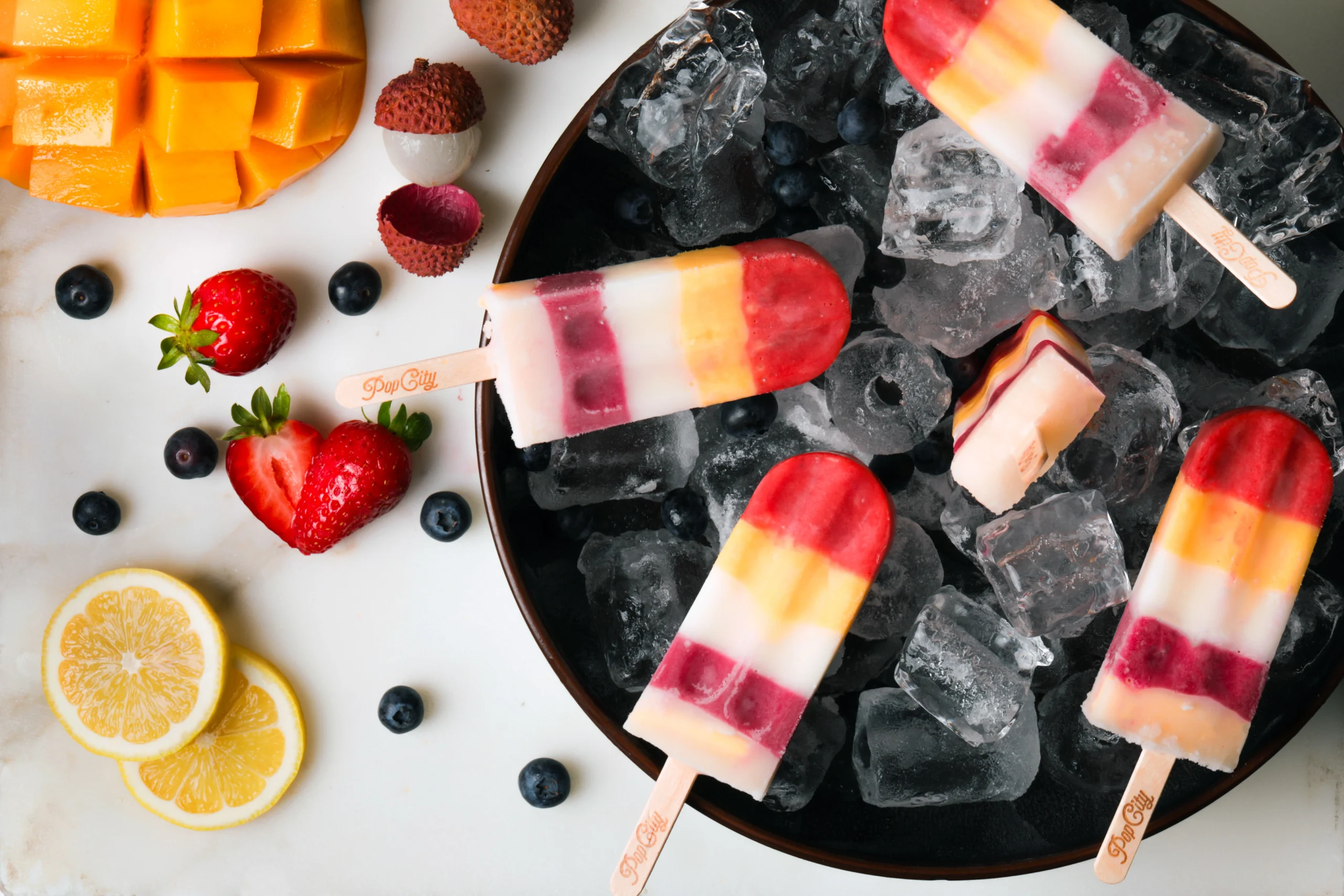 A bowl filled with vibrant, multi-layered popsicles on ice, surrounded by fresh fruits like mango, lychees, strawberries, blueberries, and lemons, labeled 'PopCity'