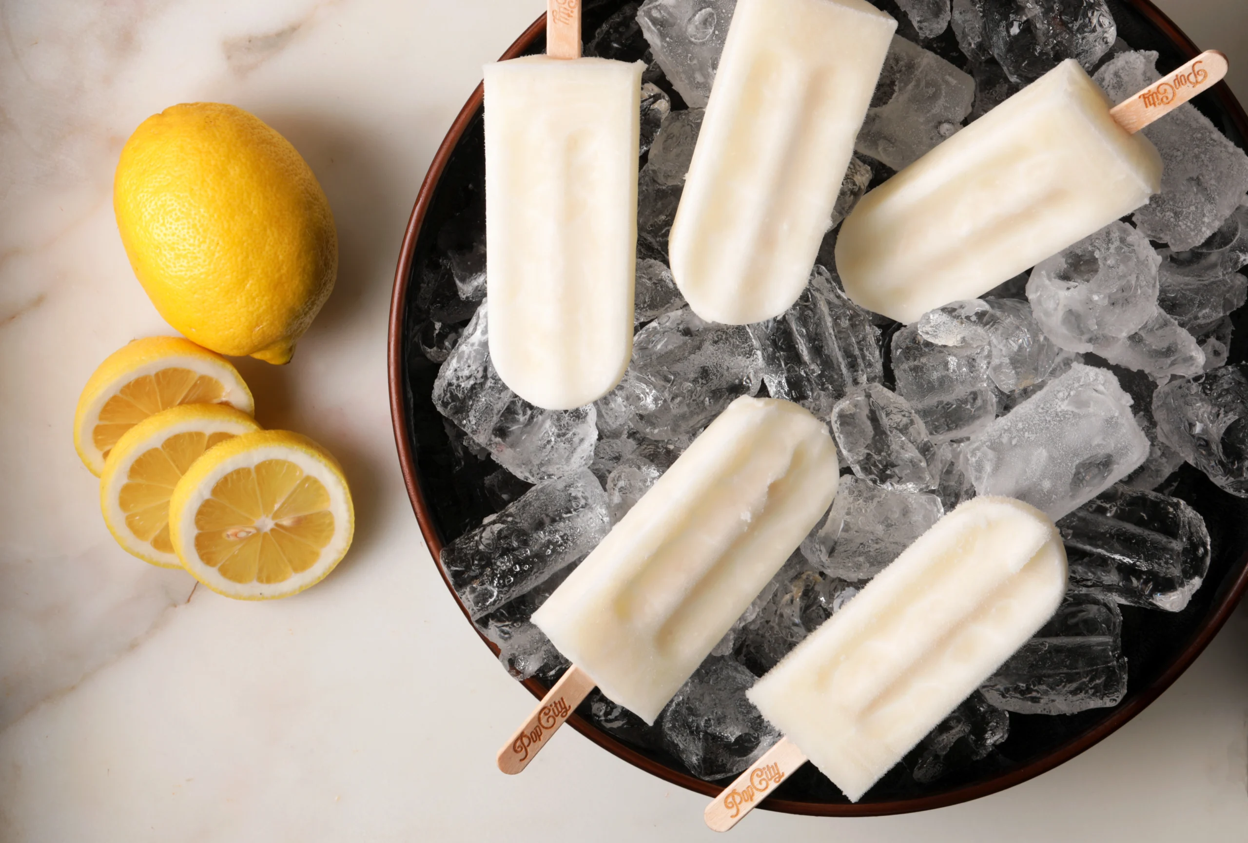 Five lemon-flavored popsicles on a bed of ice in a round container, with a whole lemon and three lemon slices on a marble surface, labeled 'PopCity'