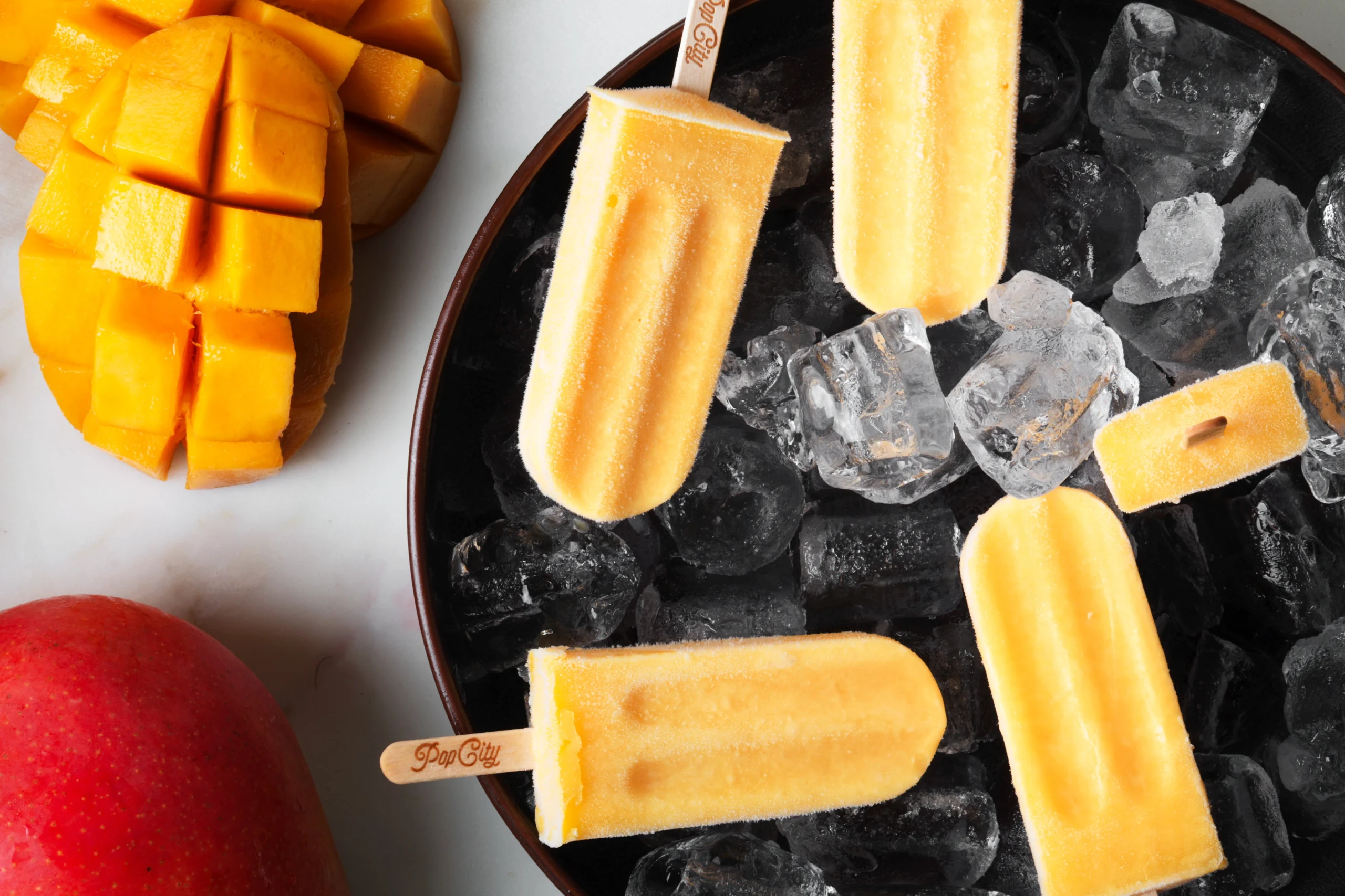 Bright yellow-orange mango popsicles on a bed of ice cubes, with sliced and whole mangoes in a dark bowl, labeled 'Pop City'