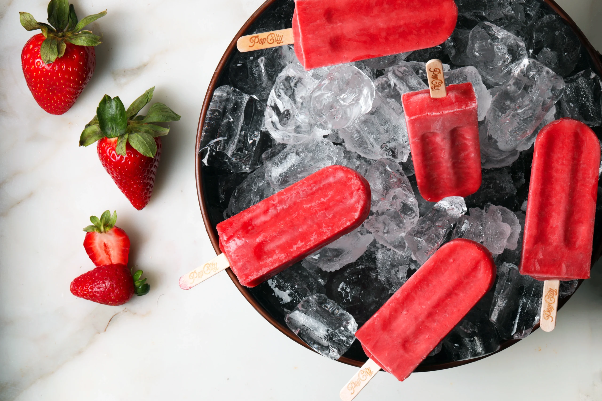 Five red strawberry popsicles in a bowl filled with ice cubes, accompanied by whole and halved strawberries, labeled 'PopCity'