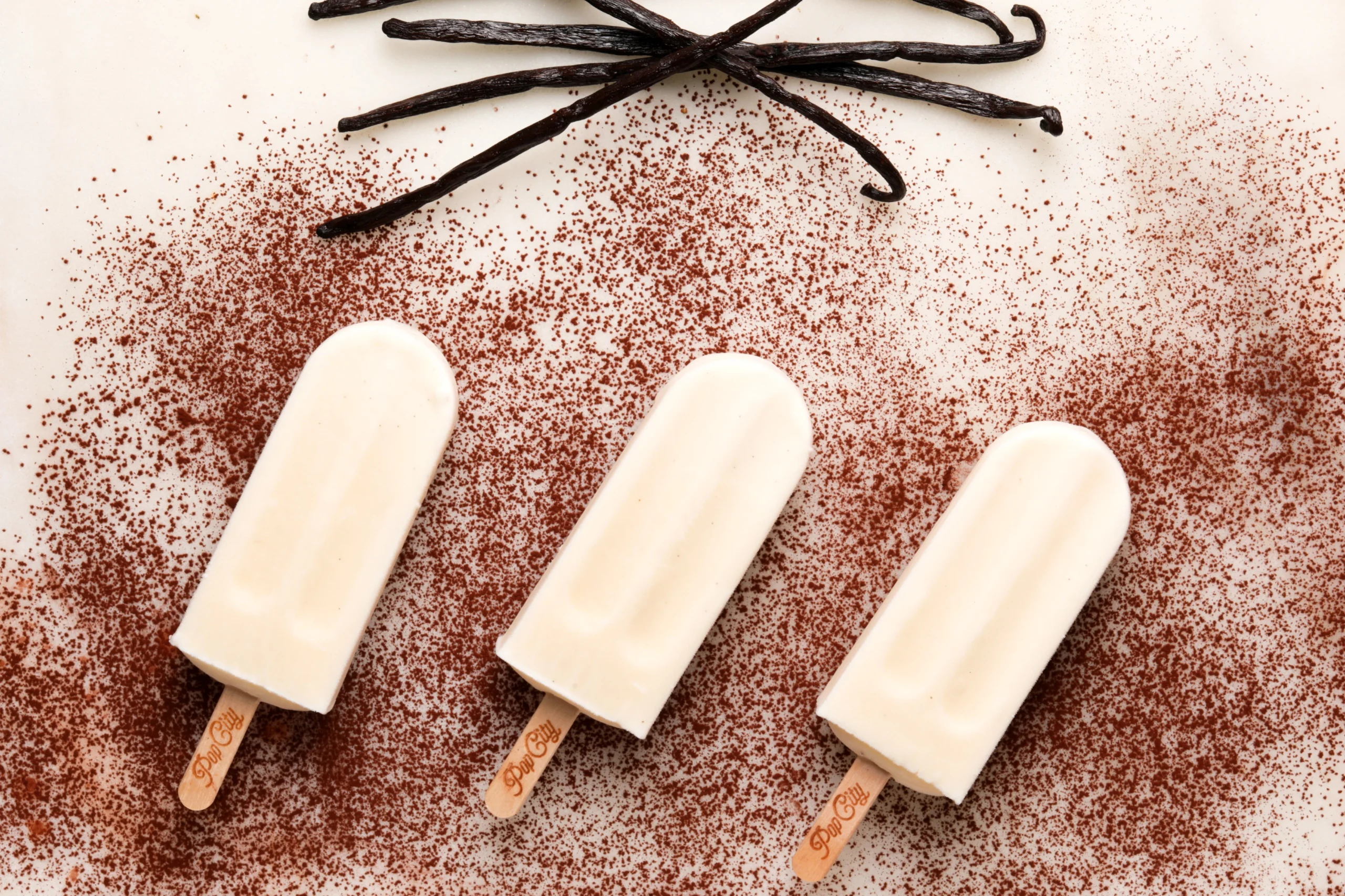 Three creamy vanilla popsicles on a cocoa-sprinkled surface, with crisscrossed vanilla beans, labeled 'PopCity'