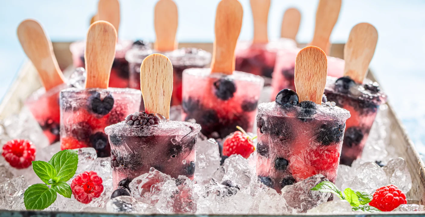 A tray filled with ice and several berry-flavored popsicles with wooden sticks, embedded with whole blueberries and raspberries, surrounded by ice cubes, fresh raspberries, and mint leaves,