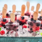 A tray filled with ice and several berry-flavored popsicles with wooden sticks, embedded with whole blueberries and raspberries, surrounded by ice cubes, fresh raspberries, and mint leaves,
