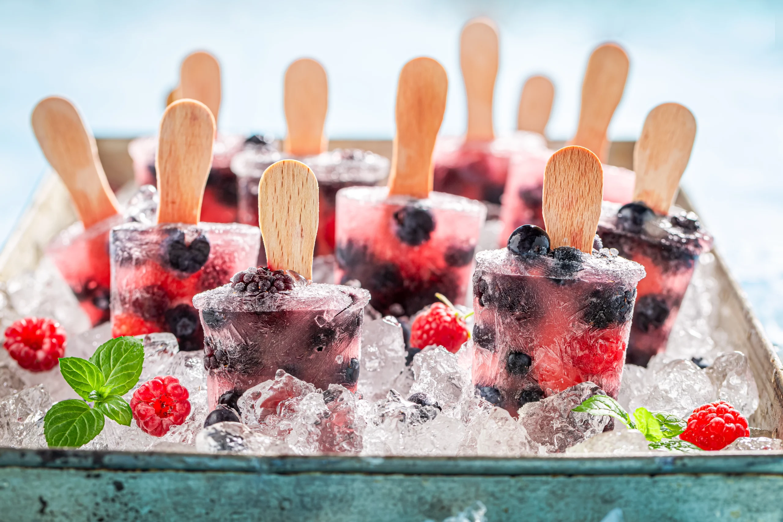 A tray filled with ice and several berry-flavored popsicles with wooden sticks, embedded with whole blueberries and raspberries, surrounded by ice cubes, fresh raspberries, and mint leaves,