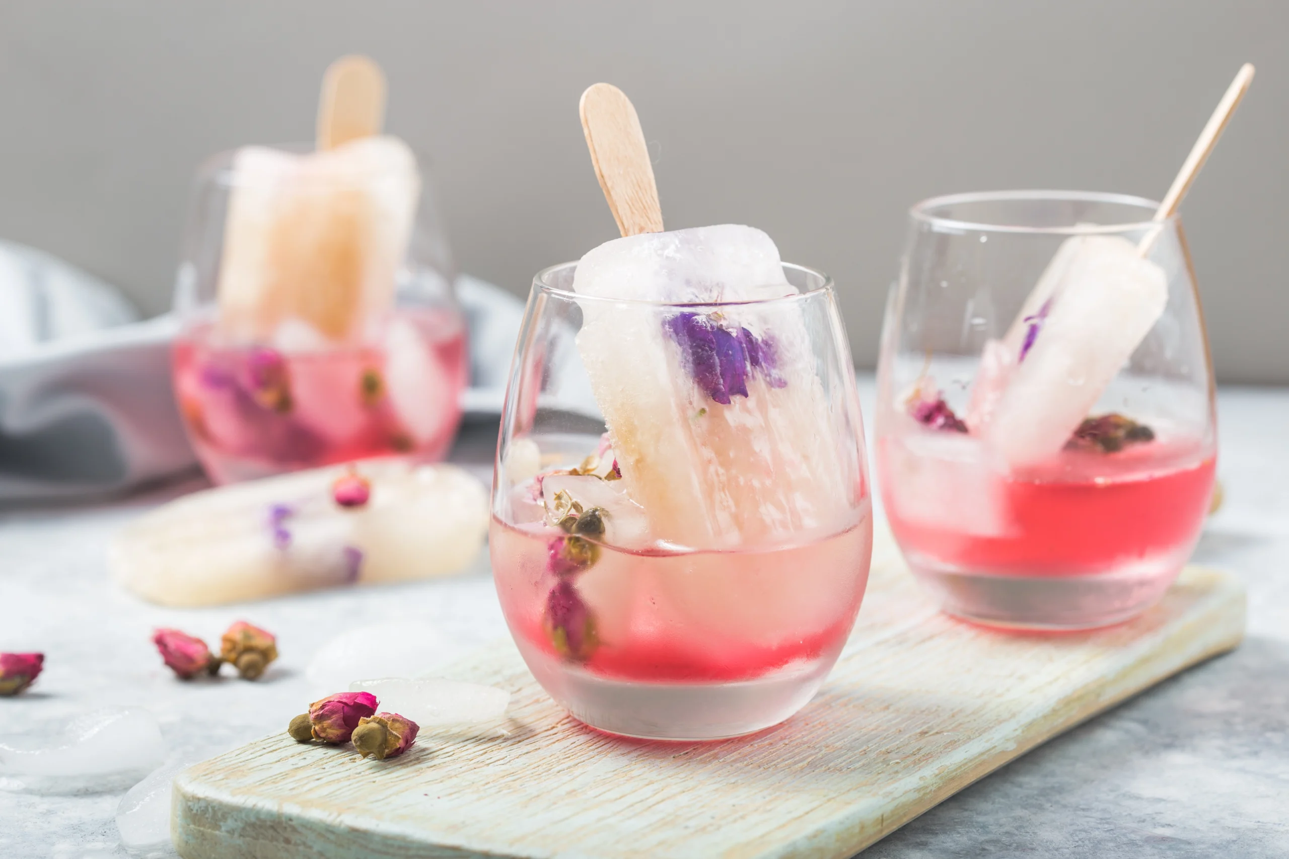 Three glasses with pink popsicles submerged in a pink-colored liquid, containing flower petals and small buds, placed on a light wooden board, with ice cubes and flower buds scattered around