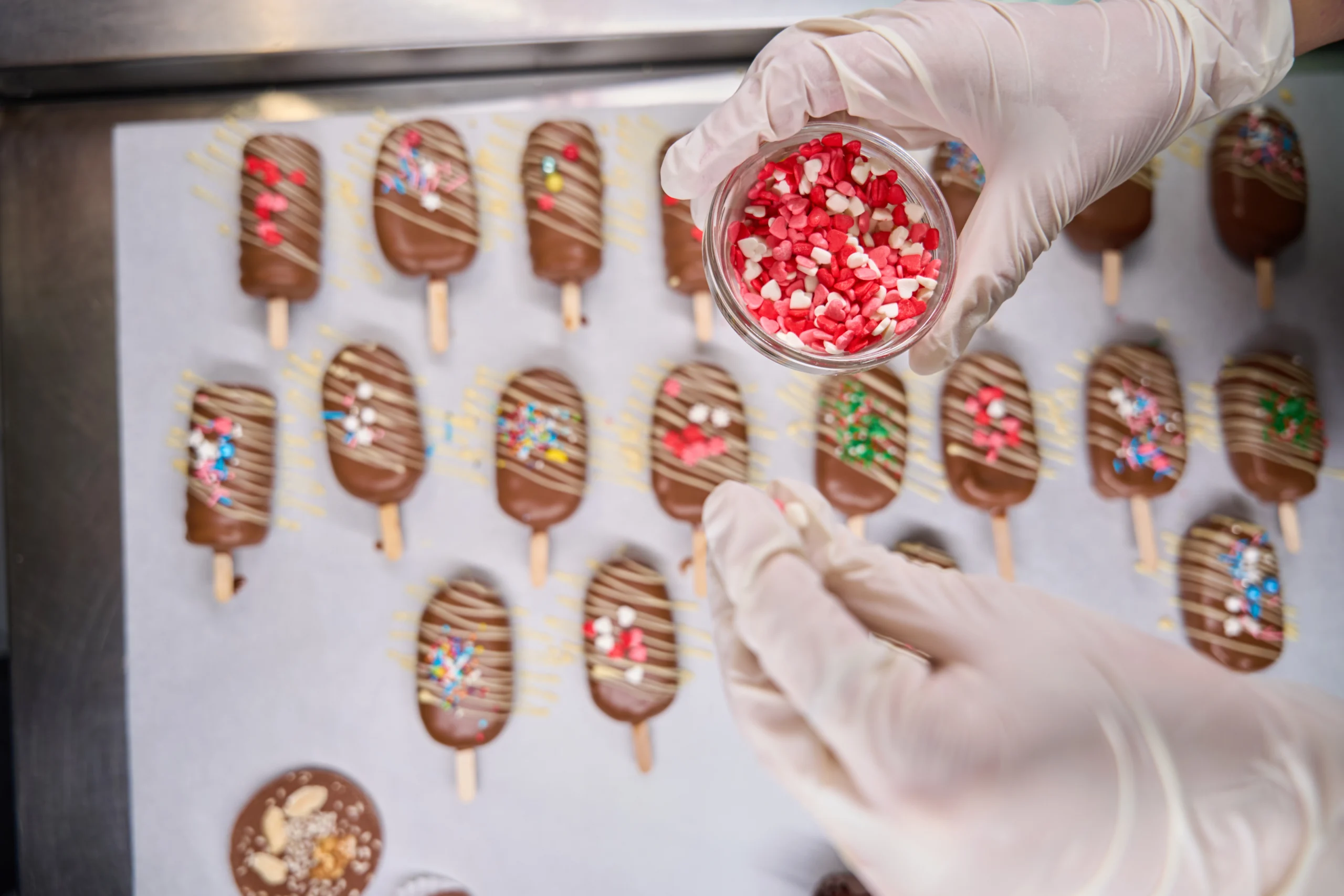 Person holding a small container filled with red and white heart-shaped sprinkles, with several chocolate-covered popsicles decorated with colorful sprinkles and drizzled with white chocolate below