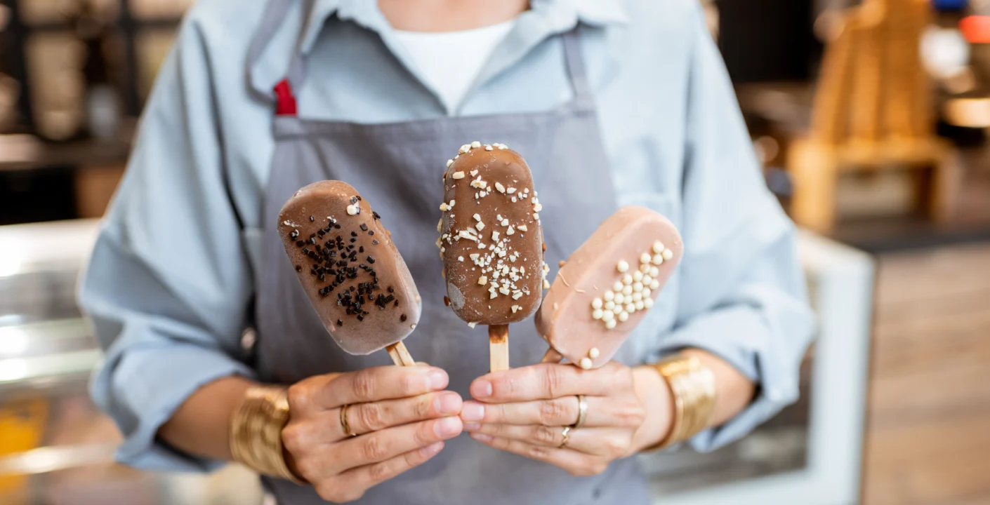 Person wearing a light blue shirt and gray apron, holding three ice cream popsicles with different chocolate coatings and sprinkles