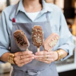 Person wearing a light blue shirt and gray apron, holding three ice cream popsicles with different chocolate coatings and sprinkles