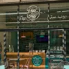 The PopCity storefront with a glass facade, showcasing its bright teal and pink branding. The store’s logo is displayed prominently above the entrance, with a branded counter inside. Teal tiles accent the walls, while pink and teal chairs add a pop of color to the outdoor seating. A promotional spinning wheel is visible inside, alongside a display case.
