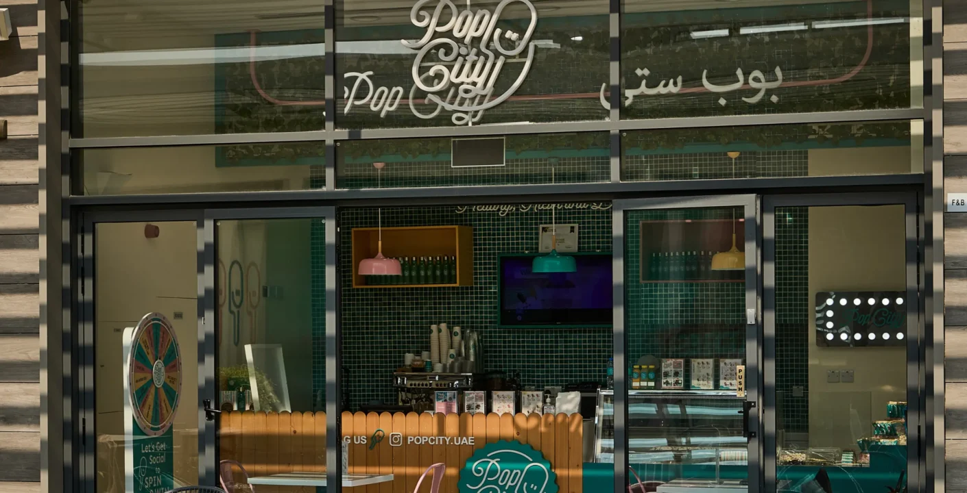 The PopCity storefront with a glass facade, showcasing its bright teal and pink branding. The store’s logo is displayed prominently above the entrance, with a branded counter inside. Teal tiles accent the walls, while pink and teal chairs add a pop of color to the outdoor seating. A promotional spinning wheel is visible inside, alongside a display case.