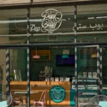 The PopCity storefront with a glass facade, showcasing its bright teal and pink branding. The store’s logo is displayed prominently above the entrance, with a branded counter inside. Teal tiles accent the walls, while pink and teal chairs add a pop of color to the outdoor seating. A promotional spinning wheel is visible inside, alongside a display case.