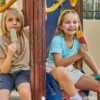Two children sitting on a playground structure, each holding and eating a popsicle. The child on the left wears a beige shirt and dark gray shorts, holding a green popsicle. The child on the right wears a light blue shirt and white shorts, holding a red and yellow popsicle. The playground includes wooden elements and colorful plastic