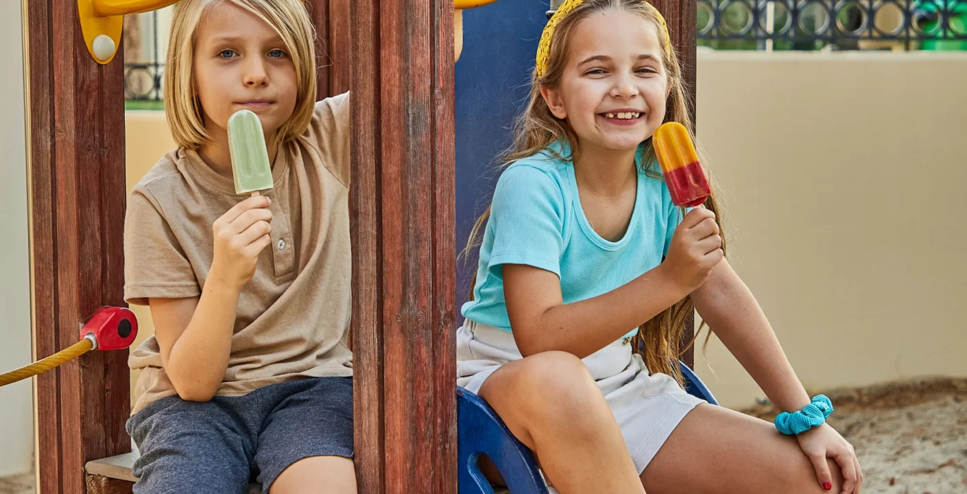Two children sitting on a playground structure, each holding and eating a popsicle. The child on the left wears a beige shirt and dark gray shorts, holding a green popsicle. The child on the right wears a light blue shirt and white shorts, holding a red and yellow popsicle. The playground includes wooden elements and colorful plastic