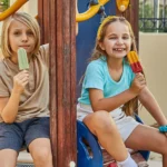 Two children sitting on a playground structure, each holding and eating a popsicle. The child on the left wears a beige shirt and dark gray shorts, holding a green popsicle. The child on the right wears a light blue shirt and white shorts, holding a red and yellow popsicle. The playground includes wooden elements and colorful plastic