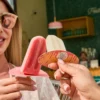 Two hands holding popsicles—one red and one white labeled against a green tiled wall with a wooden shelf of bottles and hanging lights in the background.