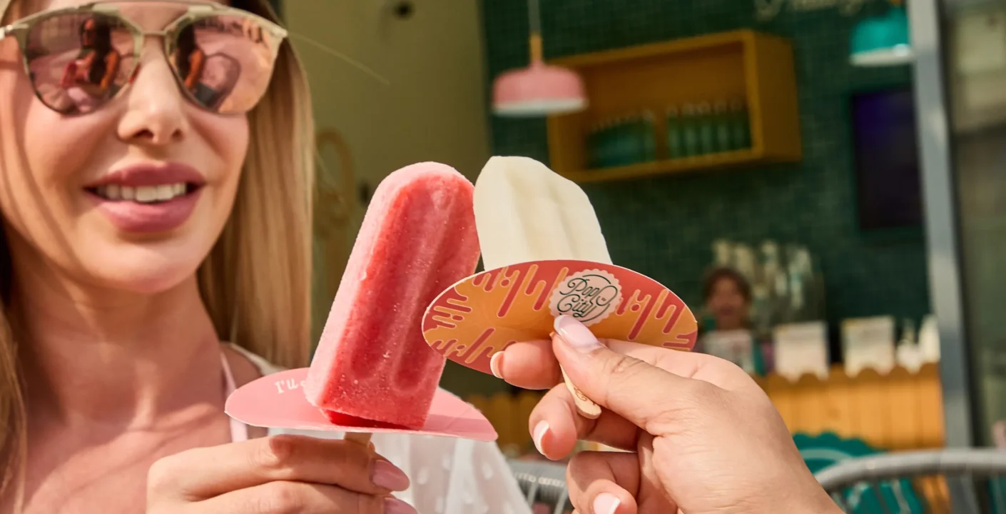 Two hands holding popsicles—one red and one white labeled against a green tiled wall with a wooden shelf of bottles and hanging lights in the background.