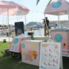 A PopCity popsicle cart with a pink and white canopy and branded umbrella, set up near a waterfront with boats in the background. A menu board displays popsicle options, with “20 AED” visible. The sunny scene includes people enjoying the area, with one person taking a photograph.