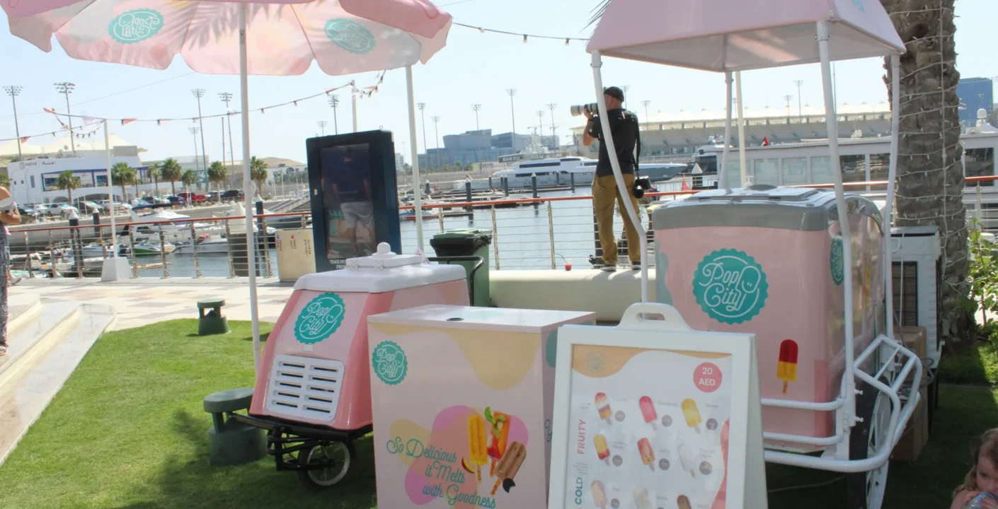 A PopCity popsicle cart with a pink and white canopy and branded umbrella, set up near a waterfront with boats in the background. A menu board displays popsicle options, with “20 AED” visible. The sunny scene includes people enjoying the area, with one person taking a photograph.