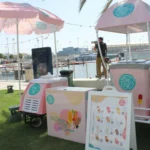 A PopCity popsicle cart with a pink and white canopy and branded umbrella, set up near a waterfront with boats in the background. A menu board displays popsicle options, with “20 AED” visible. The sunny scene includes people enjoying the area, with one person taking a photograph.