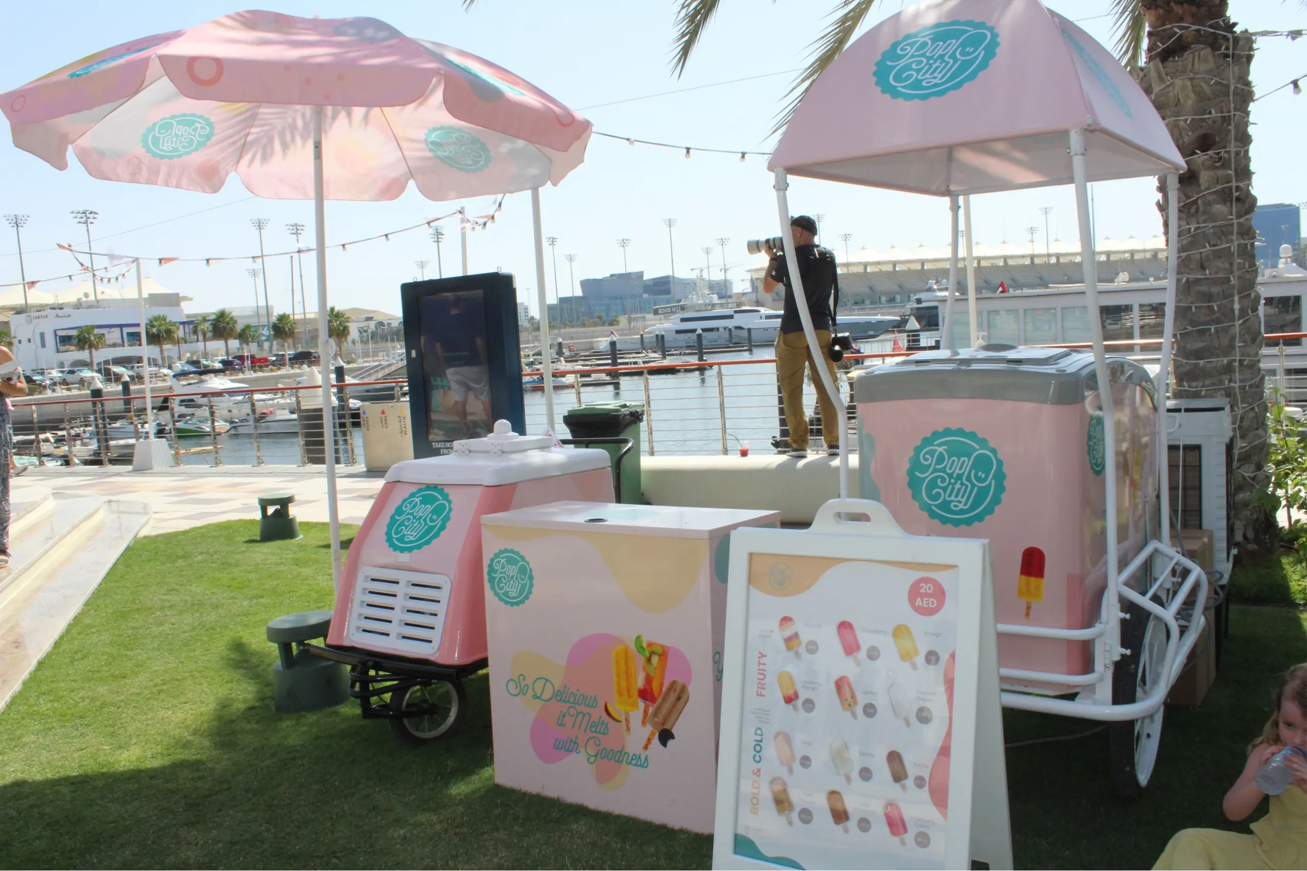 Why Pop City Pops Are the Best Dessert for Any Event in Dubai A PopCity popsicle cart with a pink and white canopy and branded umbrella, set up near a waterfront with boats in the background. A menu board displays popsicle options, with “20 AED” visible. The sunny scene includes people enjoying the area, with one person taking a photograph.