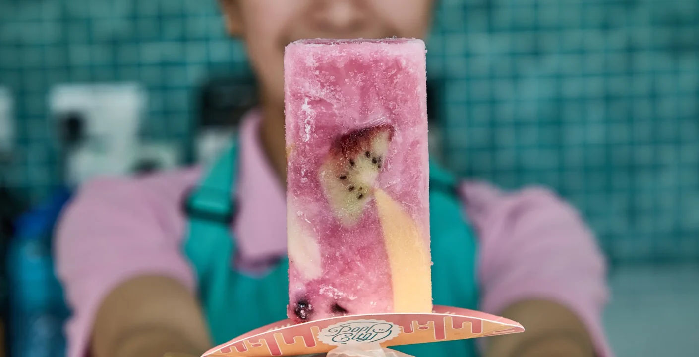 Hand holding a colorful fruit popsicle with kiwi and berries, branded Pop City stick, against a tiled wall
