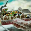 Chocolate ice cream bar with white drizzle and colorful sprinkles being decorated on a tray by a gloved hand
